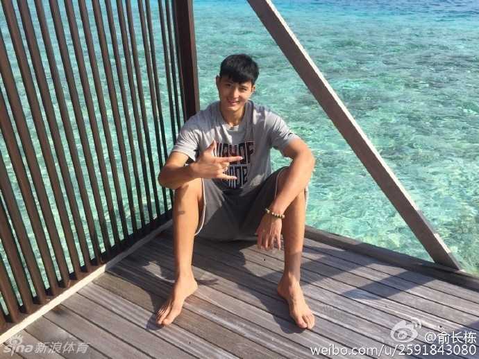 俞长栋穿着休闲夏装，在椰林树影下为妻子拍照，阳光透过树叶洒在他专注的脸上，这一刻他不是球星，只是一个记录妻子美丽的普通丈夫。