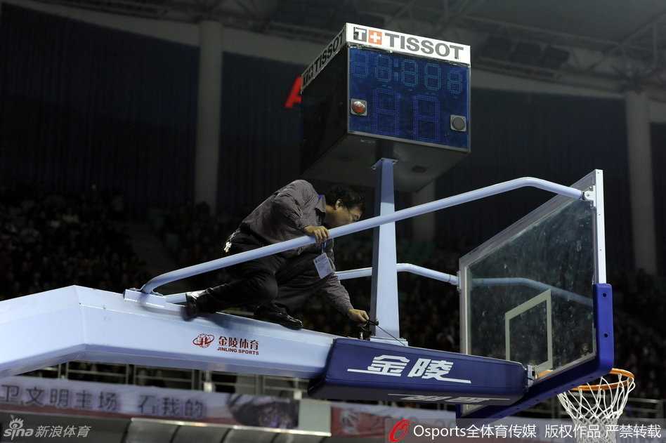 几名工作人员围聚在篮架后方，其中一人正在仔细地拨弄着计时器的线路，试图找出问题所在，场边的替补球员们也纷纷侧目关注着维修进展。