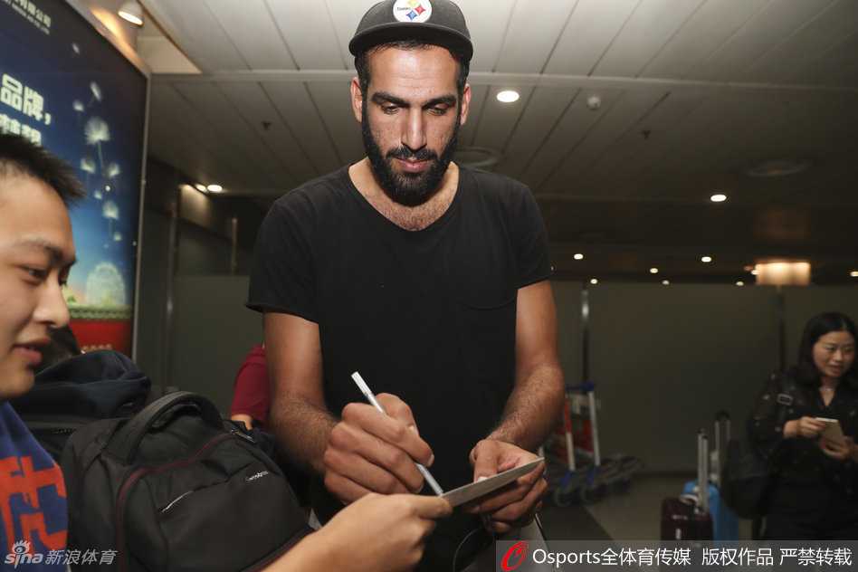 哈达迪坐在椅子上，身体微微前倾，右手握笔在球迷递来的纪念品上认真签名，侧脸的线条专注而柔和，背景是虚化的人群，这一刻他仿佛是全世界的中心。