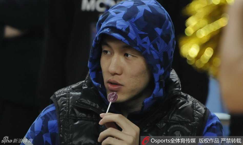 身穿长款厚实羽绒服的郭艾伦坐在场边，嘴里含着棒棒糖，眼神专注又带着一丝惬意，仿佛寒冷的天气与他无关，只有手中的甜蜜才是此刻最重要的事。