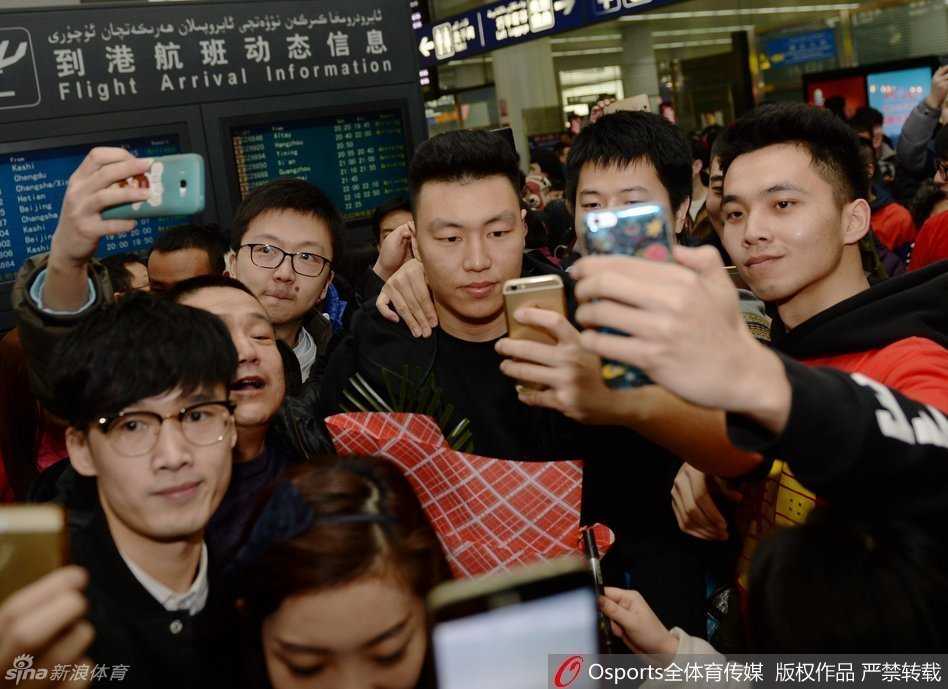 李根手捧球迷献上的鲜花，嘴角上扬露出笑容，但眼神中透露出一丝连日征战后的疲惫，身边是举起手机准备合影的兴奋球迷。