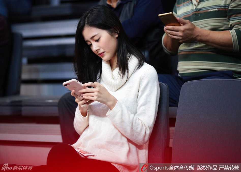 肤白如雪的女球迷在节间休息时摆弄手机，随性的穿着难掩其面容姣好，即使在昏暗的球场灯光下依然光彩照人。