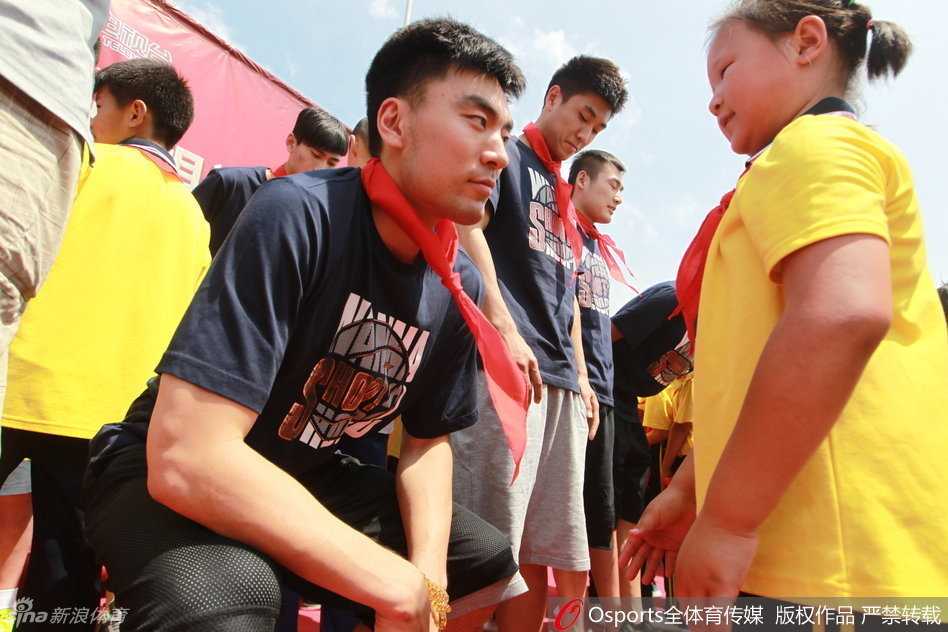 在校园操场上，辽篮队员李晓旭正与一群小学生进行篮球互动，他耐心地指导着孩子们的基础动作，画面温馨。