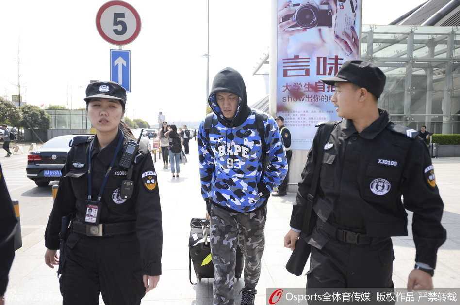 在戴着白色手套的安保人员严密的簇拥下，身穿休闲装的郭艾伦眼神坚定地走出机场通道，周围是密集的人群和闪烁的闪光灯。