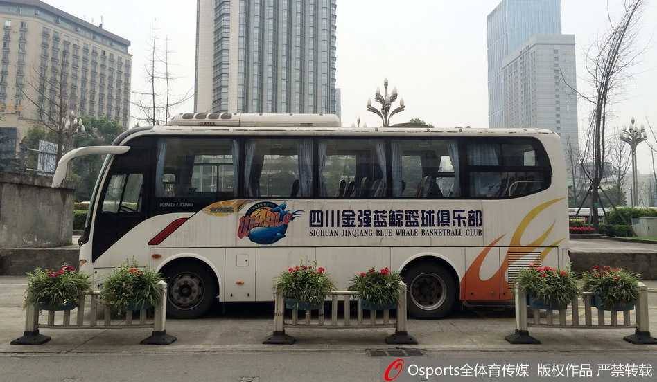 空旷的酒店门前，一辆喷涂着‘四川队’标识的蓝白大巴车孤零零地停靠在路边，车身反射着午后惨白的阳光，车内空无一人，仿佛在诉说着赛事停摆的紧张与不确定。