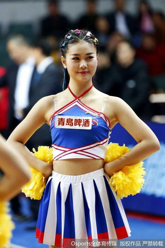 在灯光聚焦的赛场中央，一位啦啦队员正完成一个舒展的跳跃动作，曼妙的身姿定格在半空中，背后的看台上坐满了翘首以盼的观众，构成一幅动感十足的赛场画面。