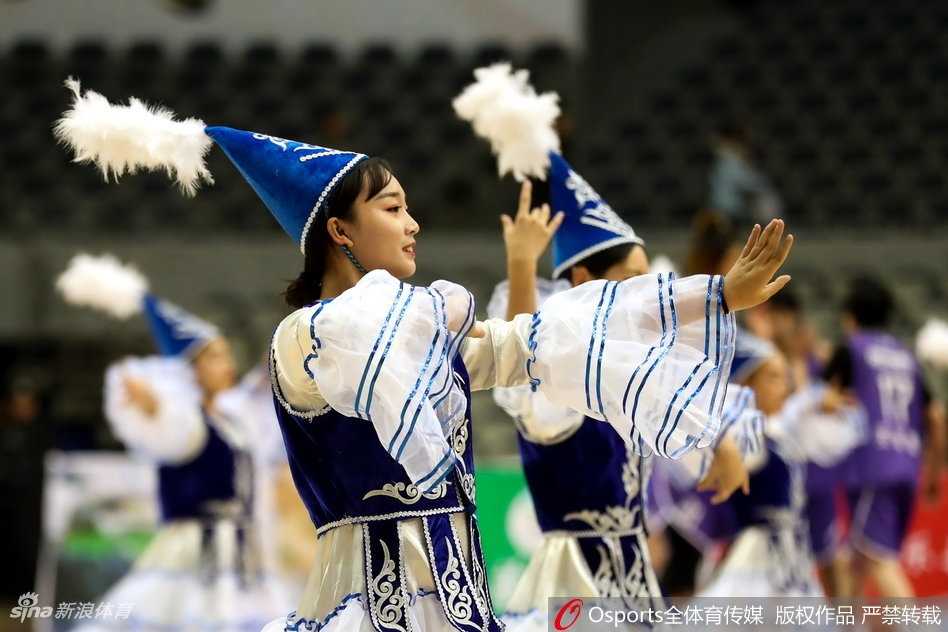 在赛场的一角，两位啦啦队员面向观众席摆出活力四射的结束pose。她们的笑容极具感染力，即使隔着屏幕也能让人感受到那份青春洋溢的热情，身后的新疆女篮队徽在灯光下熠熠生辉。