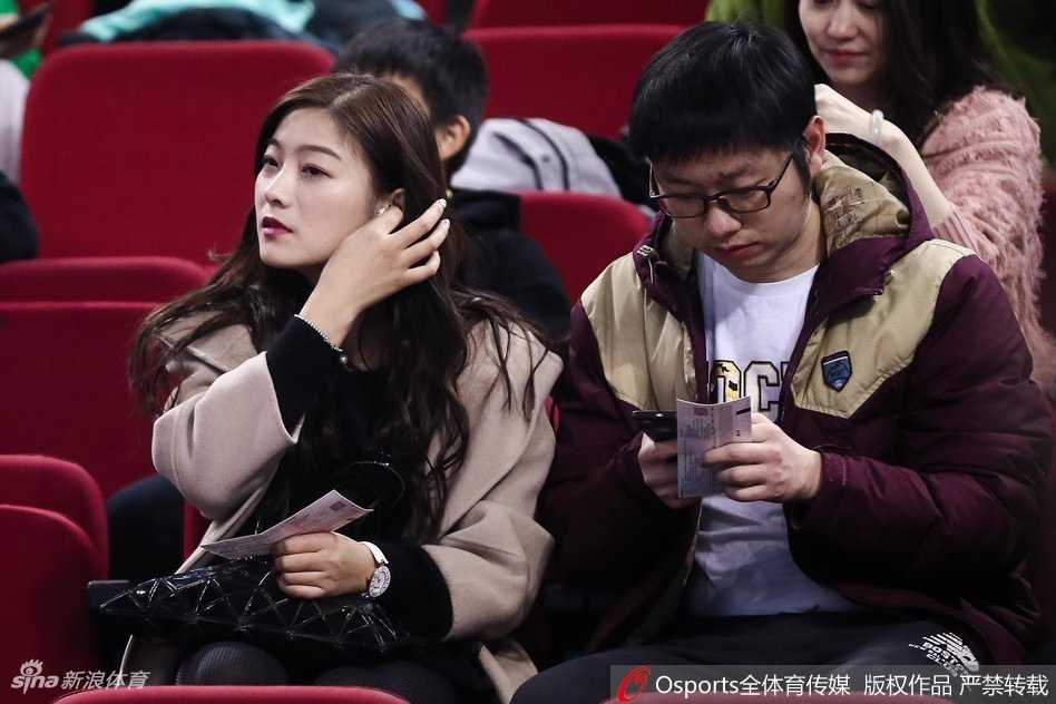 一位长发美女站在看台通道处回眸，眼神中带着一丝笑意。她身穿简约的深色外套，身后是涌动的人群和赛场明亮的灯光，整个画面充满了动态的美感，仿佛定格了比赛间隙的一个迷人瞬间。