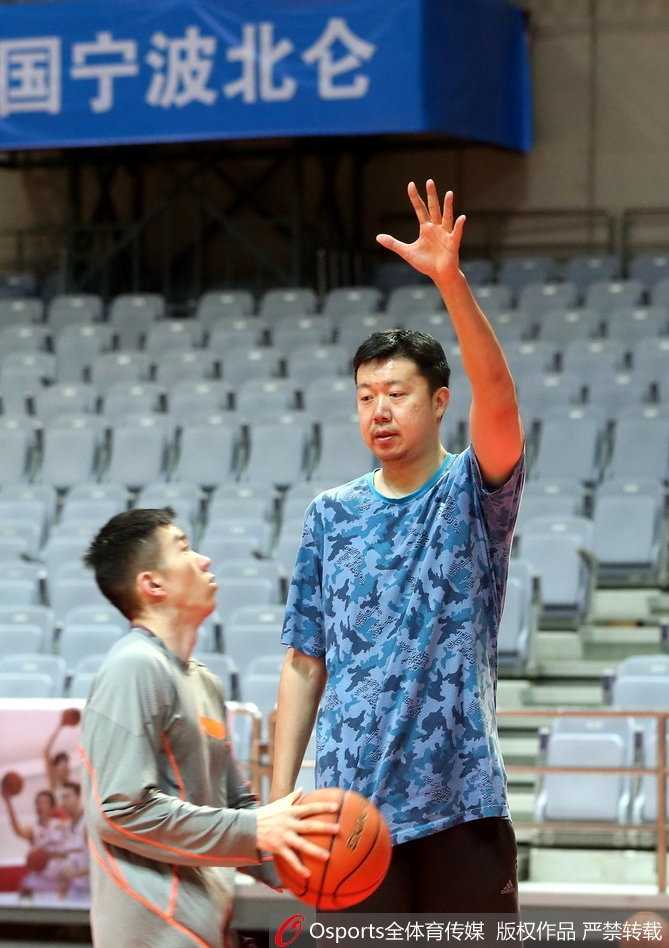 王治郅身穿深色运动外套，双手环抱胸前，目光如炬地凝视着场内球员的折返跑训练，神情专注而严肃。