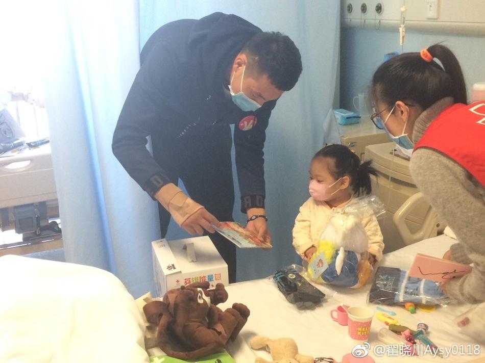 戴着口罩的小朋友坐在病床上，双手小心翼翼地打开收到的“首钢英雄能量包”，眼神中充满好奇与期待，仿佛在探寻一份神秘的宝藏。