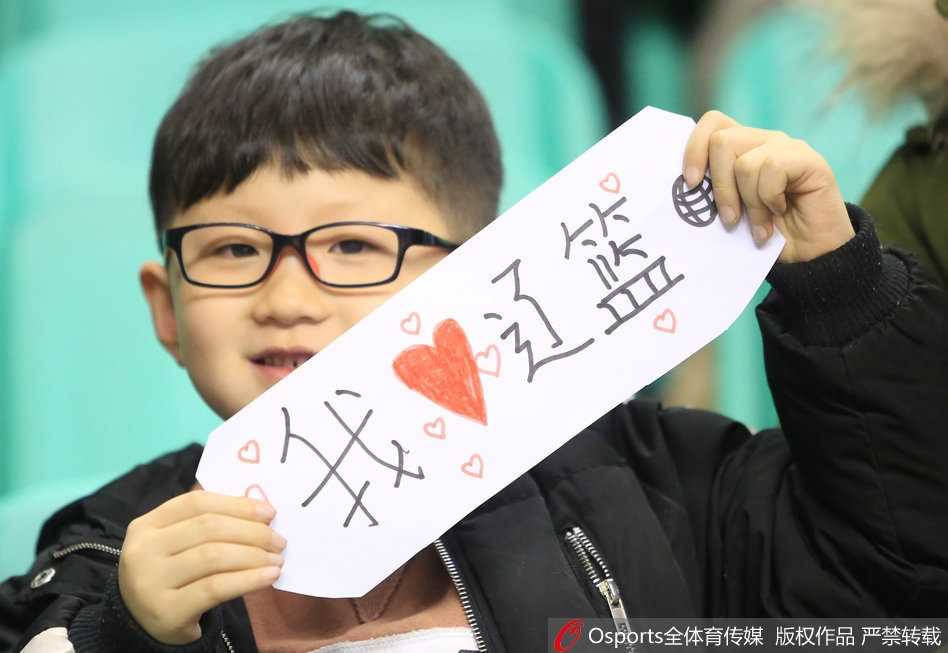 一位戴着白色毛线帽的小朋友，脸庞被冻得红扑扑的，却依然高举着自制的应援牌，上面稚嫩的笔迹写满了对辽宁男篮的喜爱，纯真的眼神紧紧追随着场内球员的一举一动。