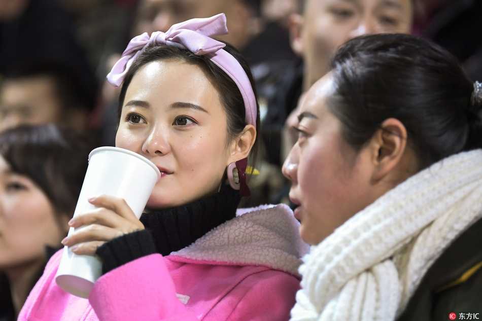 一位穿着黑色长款大衣的气质美女，双手揣在兜里，静静站立在看台过道，眼神中带着一丝疏离感，颇有大片街拍的风范，尽显高冷女神范