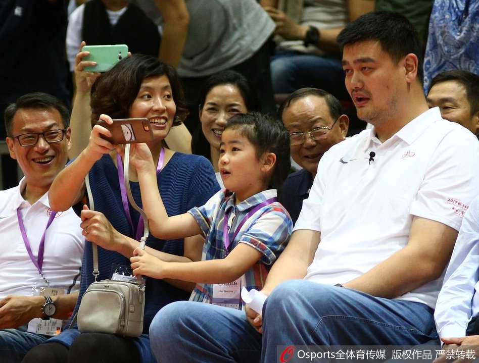 姚明身穿深色Polo衫面带微笑，目光温柔地看着场上的小球员，女儿姚沁蕾乖巧地坐在爸爸腿上，画面温馨有爱