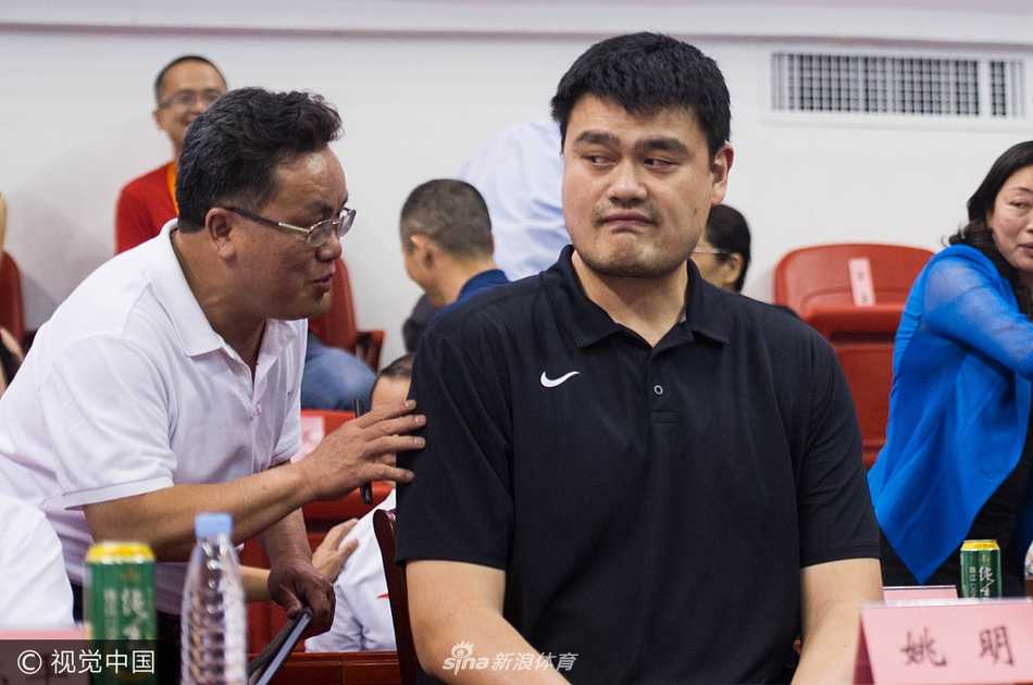 姚明现身怀集体育馆，双手自然垂放在身前，脸上带着标志性的微笑，环顾四周热情欢呼的观众，巨大的身影在灯光下显得格外亲切。