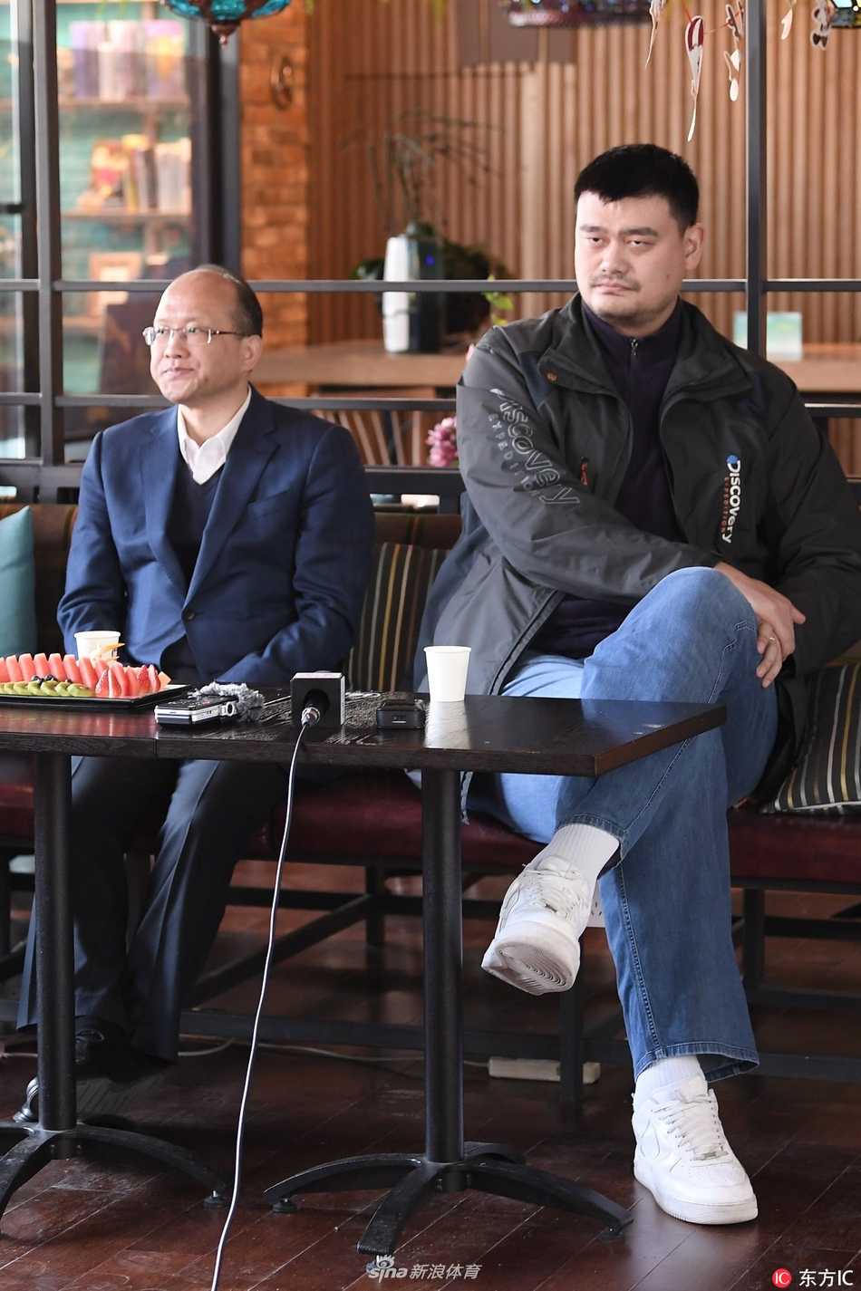 姚明身着深色休闲卫衣，双手插兜站在台侧，神情专注地聆听身旁裁判讲解技术录像，目光深邃而平和。