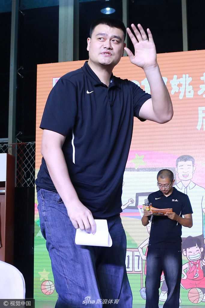 姚明将一件崭新的小号篮球服递到一名小学生手中，孩子双手接过球衣，脸上写满了惊喜与激动，身旁的嘉宾们纷纷鼓掌，姚明则低头看着孩子，眼神中充满了鼓励与期许，仿佛在说未来的赛场属于你们。