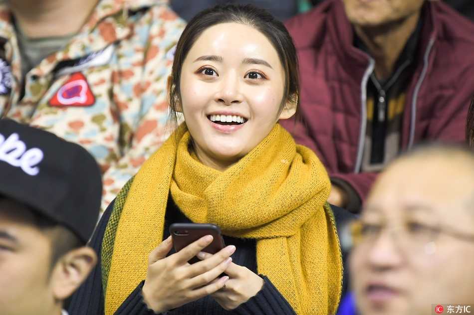 一名身穿时尚休闲装的女孩在看台上回眸，背景是欢呼雀跃的人群，她的嘴角带着淡淡的微笑，显得既冷静又迷人。