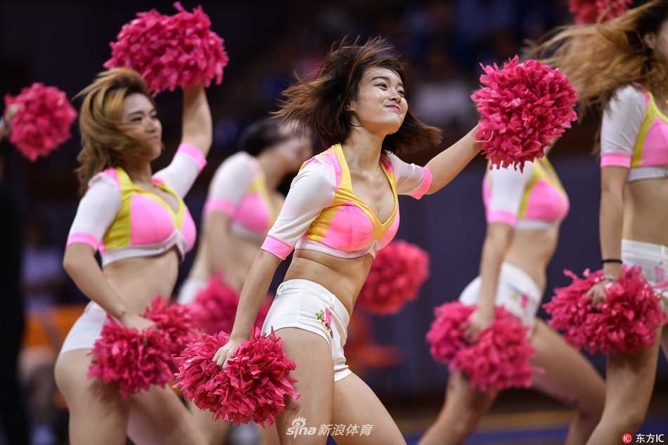 镜头聚焦于一位正在热舞的啦啦队员，她上身白色紧身衣勾勒出优美的线条，下身蓝色热裤格外亮眼。她面带自信的微笑，正随着音乐节奏摆动手臂，身后的队友们形成了整齐的队列。