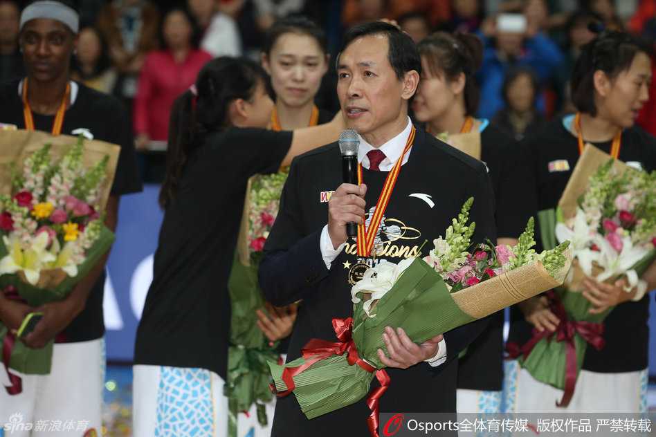 北京女篮全体成员在夺冠后于场地中央拍摄全家福，队员们有的抱着花束，有的高举着象征荣誉的手指，每个人的脸上都洋溢着幸福满足的微笑。