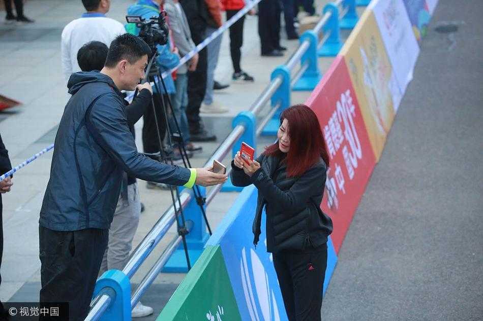 莞马赛场边的温情一幕:冬日娜与杜锋互加好友 冬日娜身披央视外套,微笑着拿出手机,杜锋则弯下腰凑近屏幕,两人在喧闹的马拉松赛场边专注地扫描二维码,背景是模糊的人群和赛事横幅,画面充满了人情味。