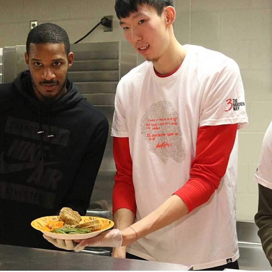 融入·感恩:周琦与哈登阿里扎的休城温情时刻 周琦与阿里扎手持餐盘,对着镜头做出搞怪的表情,两人身后是摆放整齐的节日餐点,整个画面充满了队友间的默契与欢乐。