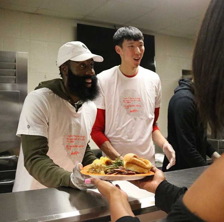 融入·感恩:周琦与哈登阿里扎的休城温情时刻 在摆满餐盘的桌子前,哈登露出标志性的大胡子笑容,耐心地为民众服务,周琦在一旁协助,画面充满了社区大家庭的温馨感。