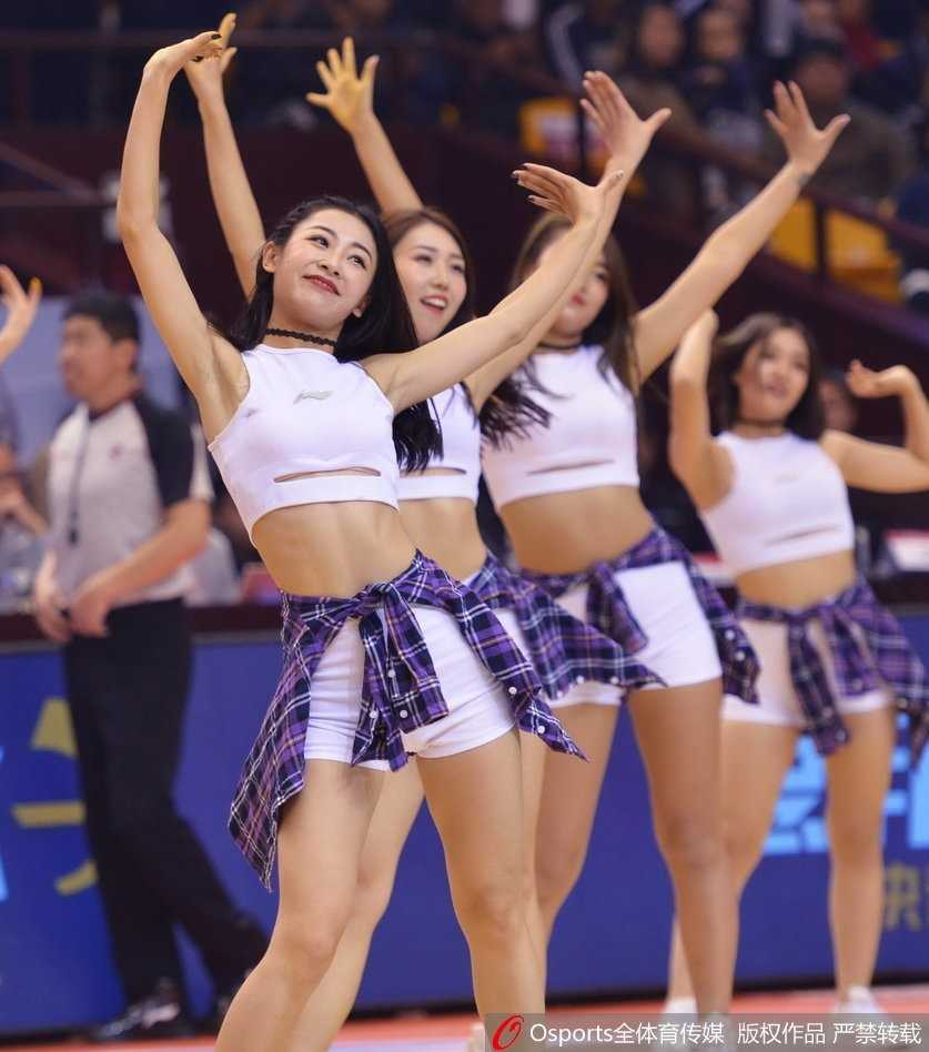 三位篮球宝贝背对镜头，展现出整齐划一的舞蹈动作和充满活力的背影，她们修长的身姿与球场地面反射的倒影相映成趣，构成一幅动感十足的画面