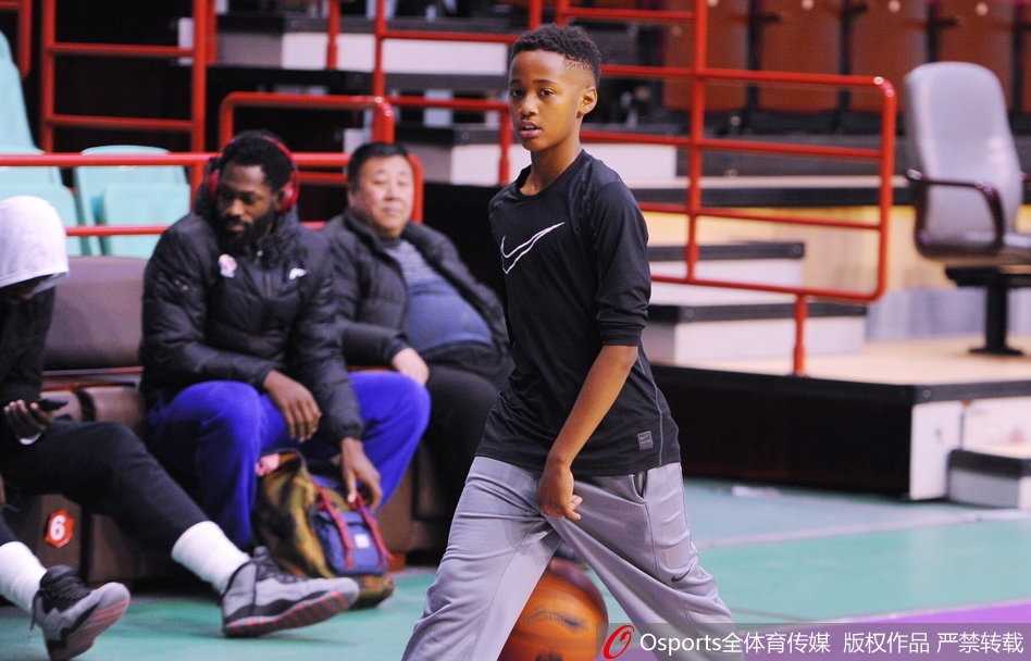 巴斯儿子双手举着篮球准备投篮，动作有模有样，巴斯在场边微笑观看