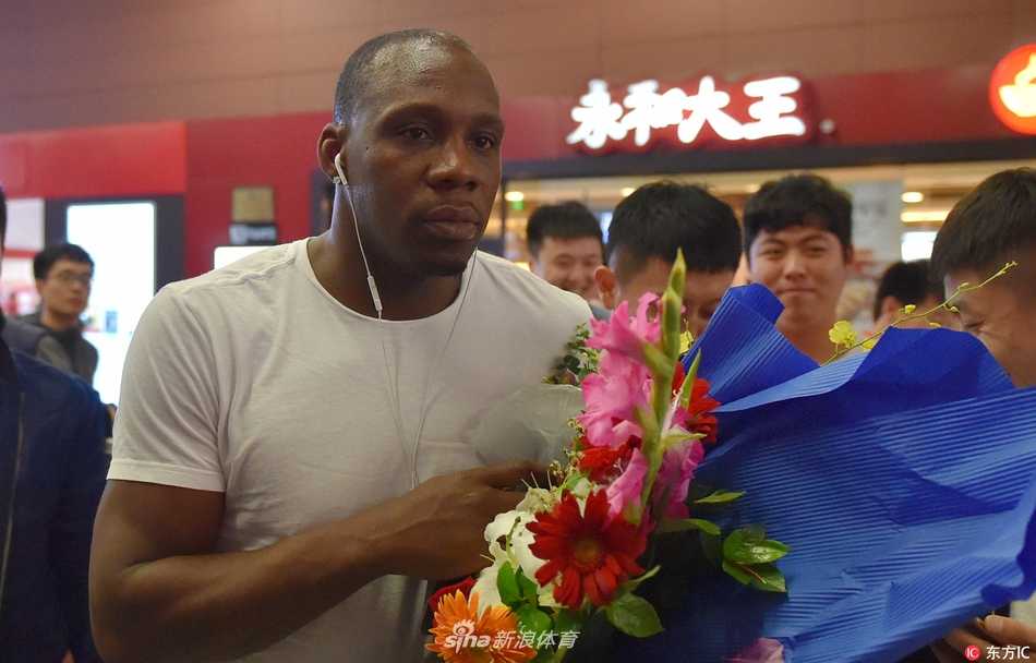 哈德森手推行李车走出通道，目光坚定地望向远方，身旁簇拥着满脸笑容的球迷，背景里隐约可见“欢迎回家”的横幅，氛围热烈而有序。