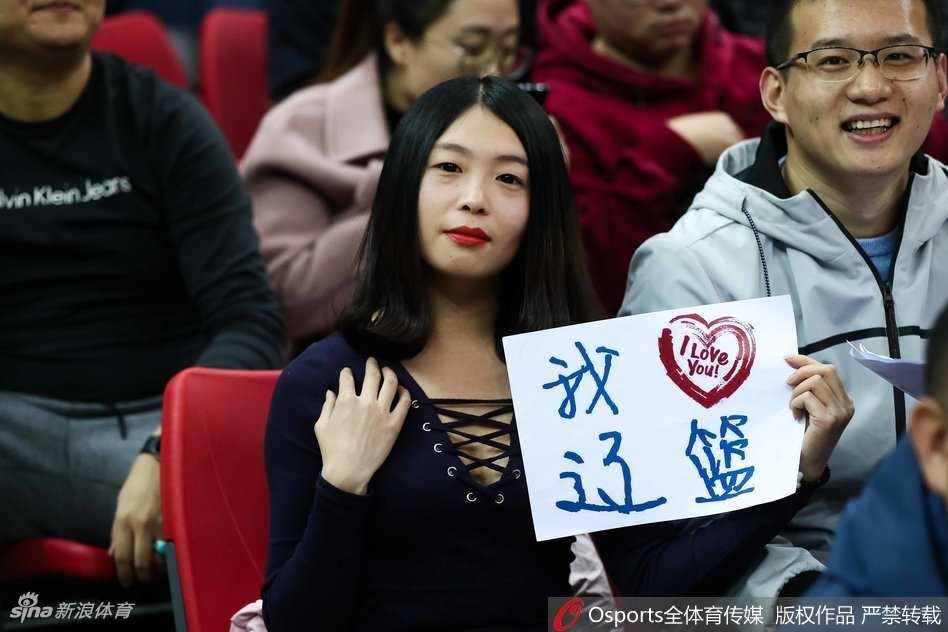 远征的赤木晴子:辽宁男篮的美女球迷客场闪耀记 一位气质文静的辽宁美女球迷,面带微笑坐在看台上,身后是喧嚣的人群,她的笑容为紧张的比赛增添了一抹亮色。
